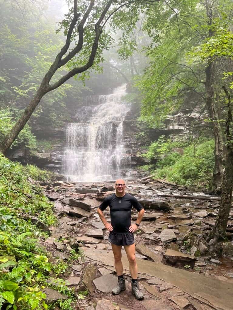 Hiker standing at the base of Ganoga Falls in Ricketts Glen State Park, with the multi-tiered waterfall rising behind him.