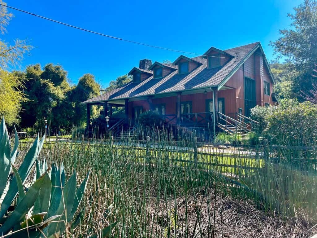 The historic Daley Ranch house surrounded by tall grasses and agave plants, with a wooden porch and green trim set against oak trees and a bright blue sky in Escondido, California.