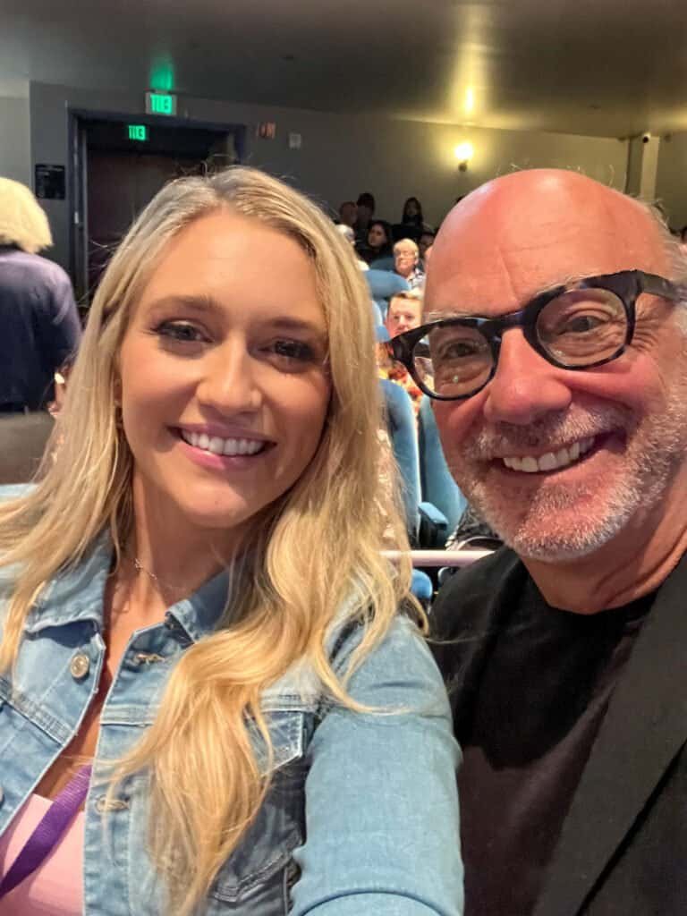 Smiling audience members seated inside a theater during a Wheel of Fortune contestant audition event, taken as a close-up selfie.