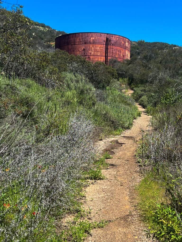 Narrow dirt section of the Sage Trail at Daley Ranch leading uphill toward a large rusted water tank surrounded by dense chaparral under a clear blue sky.