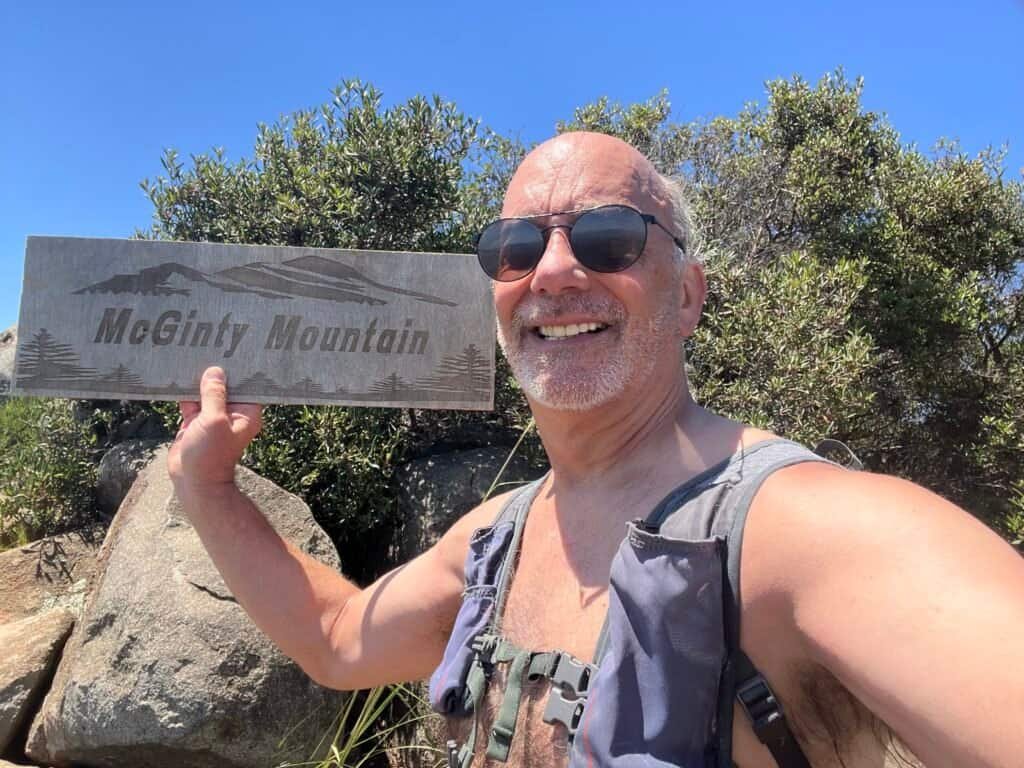 Hiker smiling at the summit of McGinty Mountain holding the wooden summit sign on a sunny day.