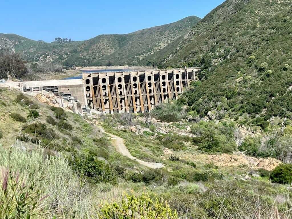 Wide view of Lake Hodges Dam with arched concrete structure, green hillsides, and a dirt trail leading into the San Dieguito Gorge
