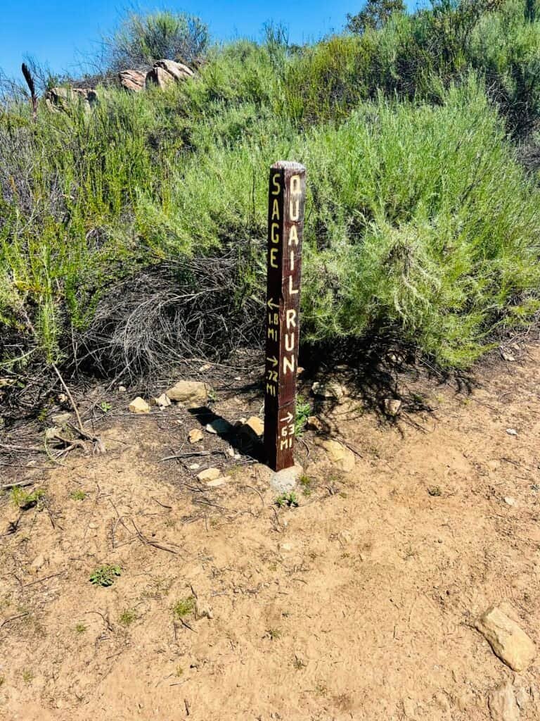 Trail post marking the junction of Sage Trail and Quail Run Trail at Daley Ranch, with scrub brush and open terrain in the background.