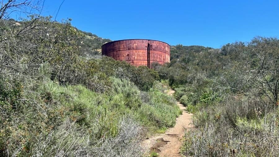 Narrow dirt trail winding uphill toward an old rusted water tank at Daley Ranch in Escondido, surrounded by chaparral and rolling hills.