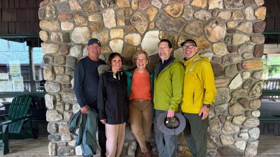 A group photo taken in front of a large stone fireplace at Adirondack Loj. From left to right: John, Yasamin, Vicki, Steve S, and Steve A, all dressed in rain gear after the Mt. Marcy hike.