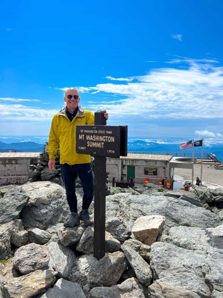 Stephen standing at the Mount Washington summit sign under blue skies