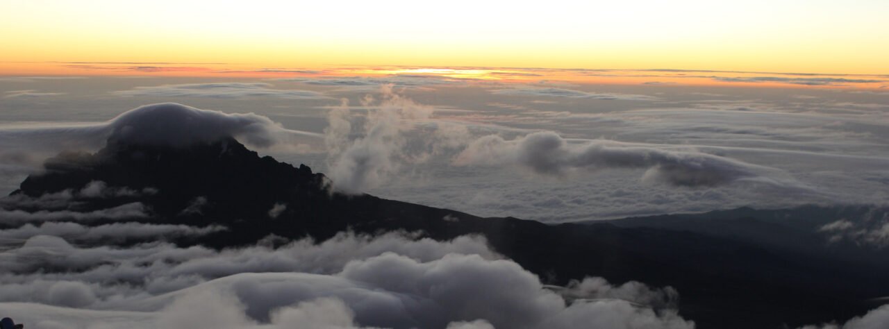 Sunrise from near the summit of Mount Kilimanjaro, with golden light spreading above a sea of clouds, a dark ridgeline silhouetted in the distance, and icy glacier formations in the foreground.