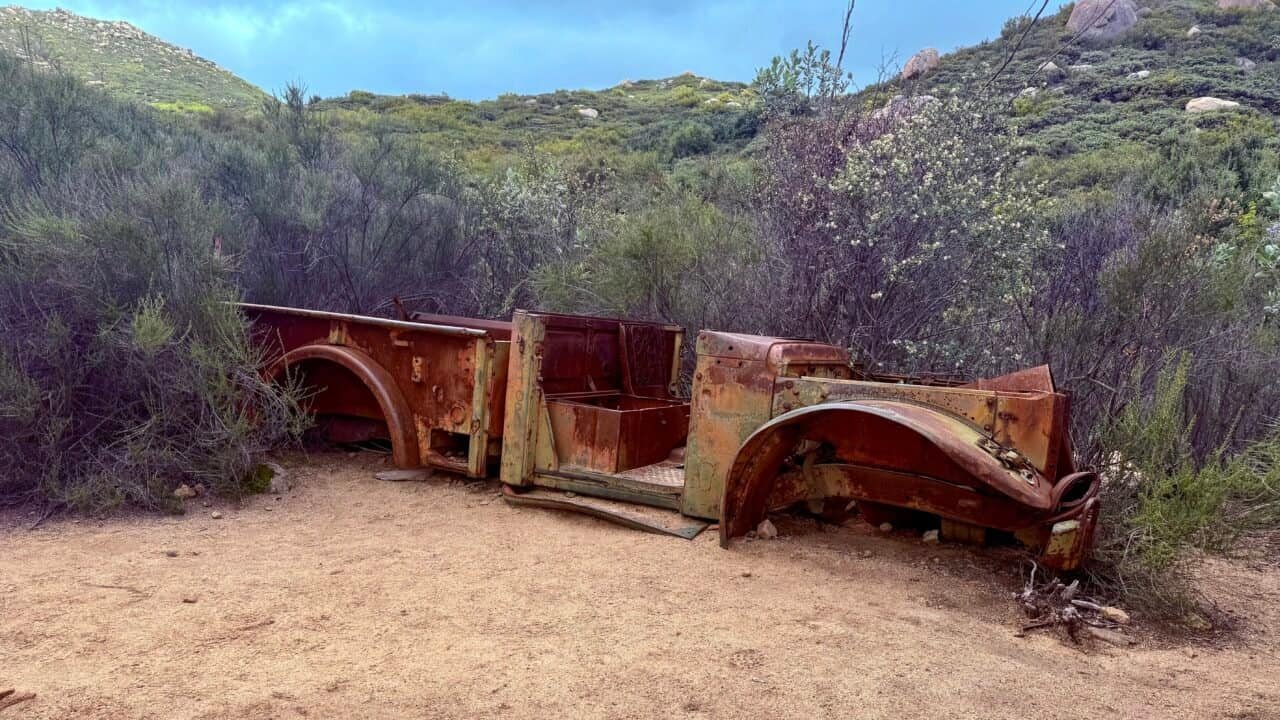 Rusted remnants of an old vehicle frame lying along the El Cajon Mountain trail in Lakeside, California, surrounded by chaparral and hillside vegetation.