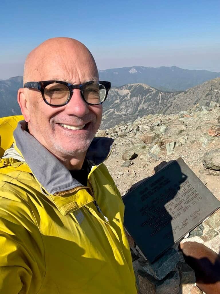 Hiker smiling beside the Wheeler Peak summit plaque at 13,161 feet, marking the highest point in New Mexico.