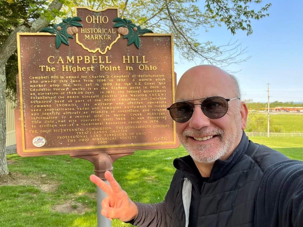 Visitor standing beside the Ohio Historical Marker at Campbell Hill, pointing to the sign marking the highest point in Ohio