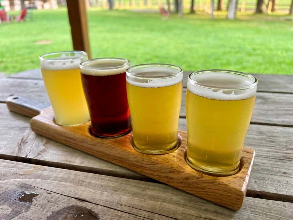 Flight of craft beers served on a wooden paddle at Ledge Brewing Company with outdoor seating and lawn in the background.