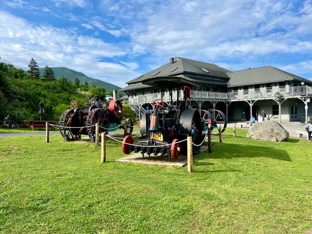 View of the Mount Washington Cog Railway base area with tracks, grassy grounds, and the White Mountains in the distance under dramatic skies.