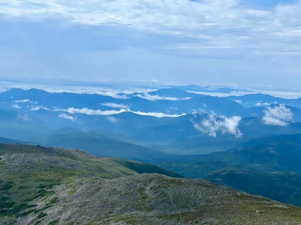 Wide summit view from Mount Washington showing layered White Mountain ridgelines and clouds drifting through the valleys below.