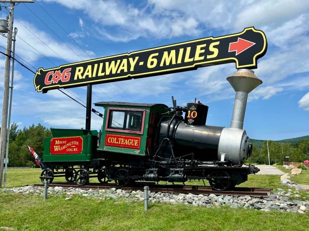 Historic Mount Washington Cog Railway steam locomotive displayed beneath a sign reading “Cog Railway – 6 Miles” in New Hampshire.