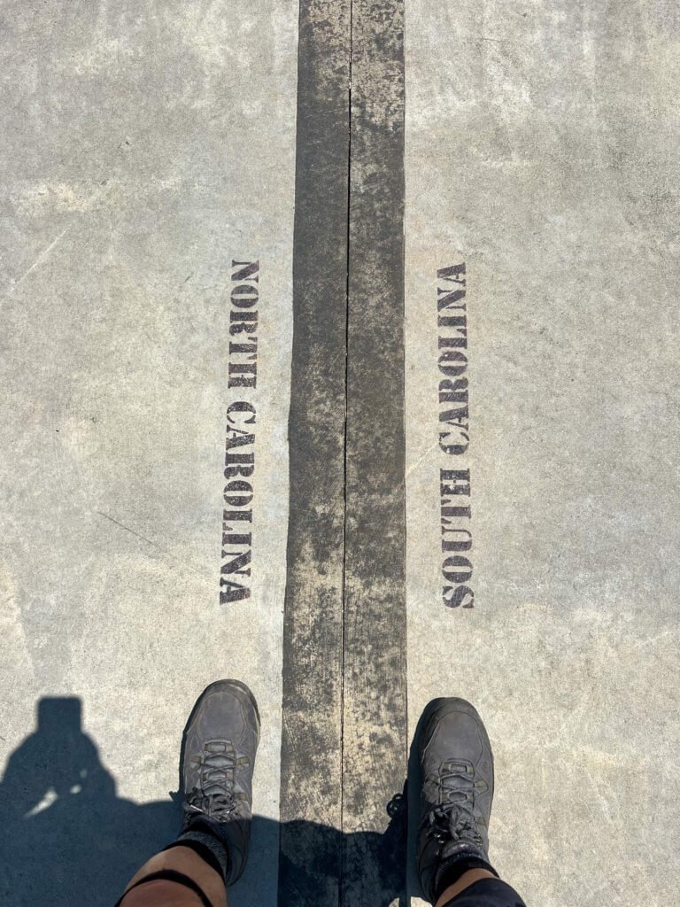Hiker standing with one foot in North Carolina and one foot in South Carolina on the state line at Sassafras Mountain