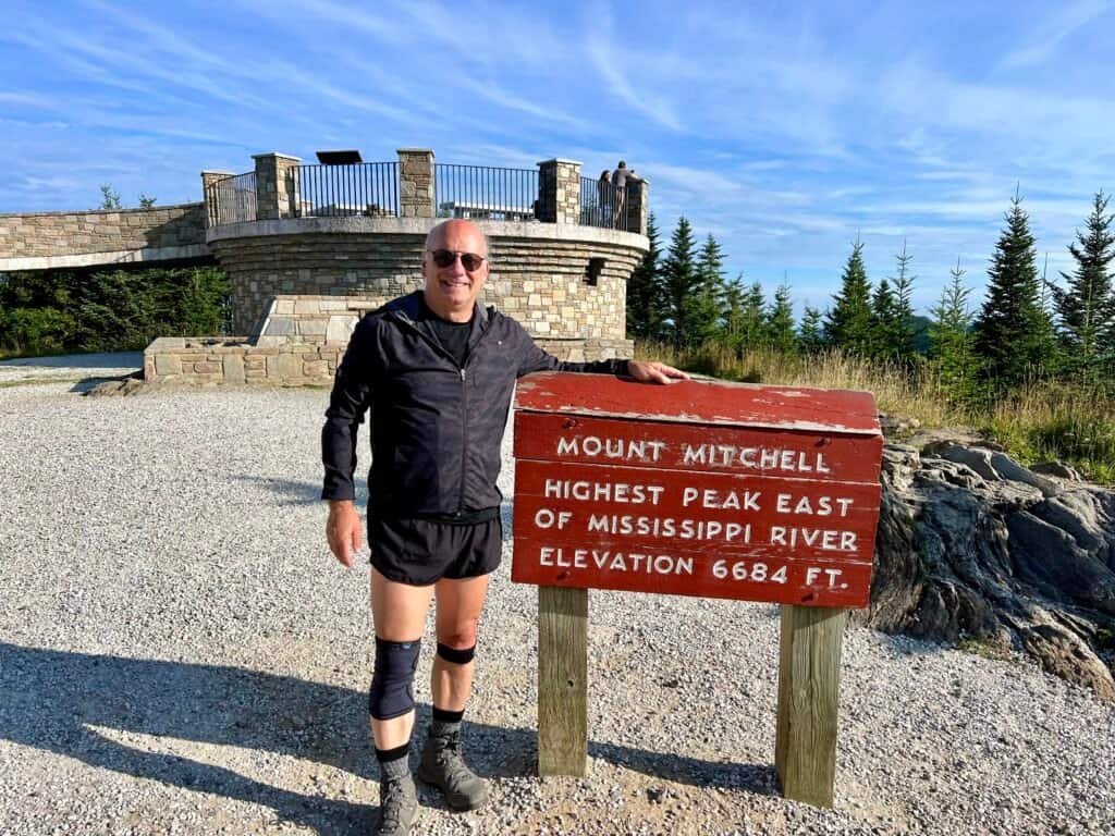Hiker standing at the Mount Mitchell summit sign near the observation tower
