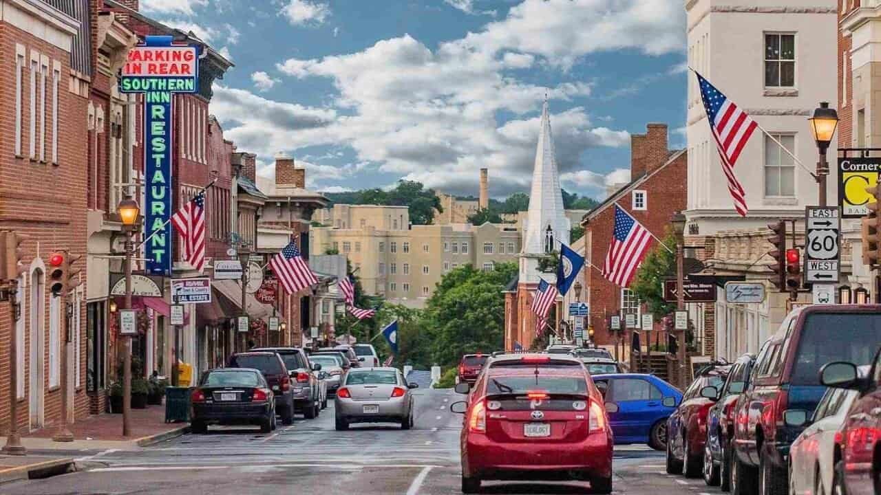 Downtown Lexington, Virginia with brick storefronts, American flags lining Main Street, and a church steeple in the distance.