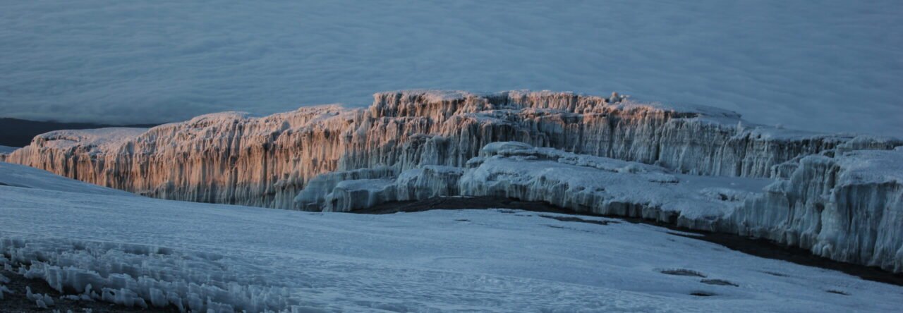 Sunrise illuminating the summit glacier on Mount Kilimanjaro, with ice formations in the foreground and a sea of clouds in the background.