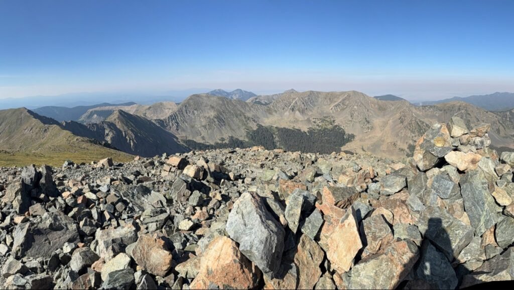 Panoramic view from the summit of Wheeler Peak, showing rocky terrain in the foreground and endless ranges of the Sangre de Cristo Mountains under a clear blue sky.