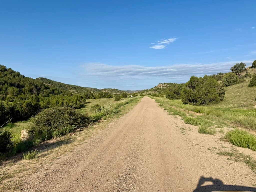 A long dirt road stretches through rolling green hills and scattered shrubs under a clear blue sky, showing the remote ranchland terrain leading toward Black Mesa.