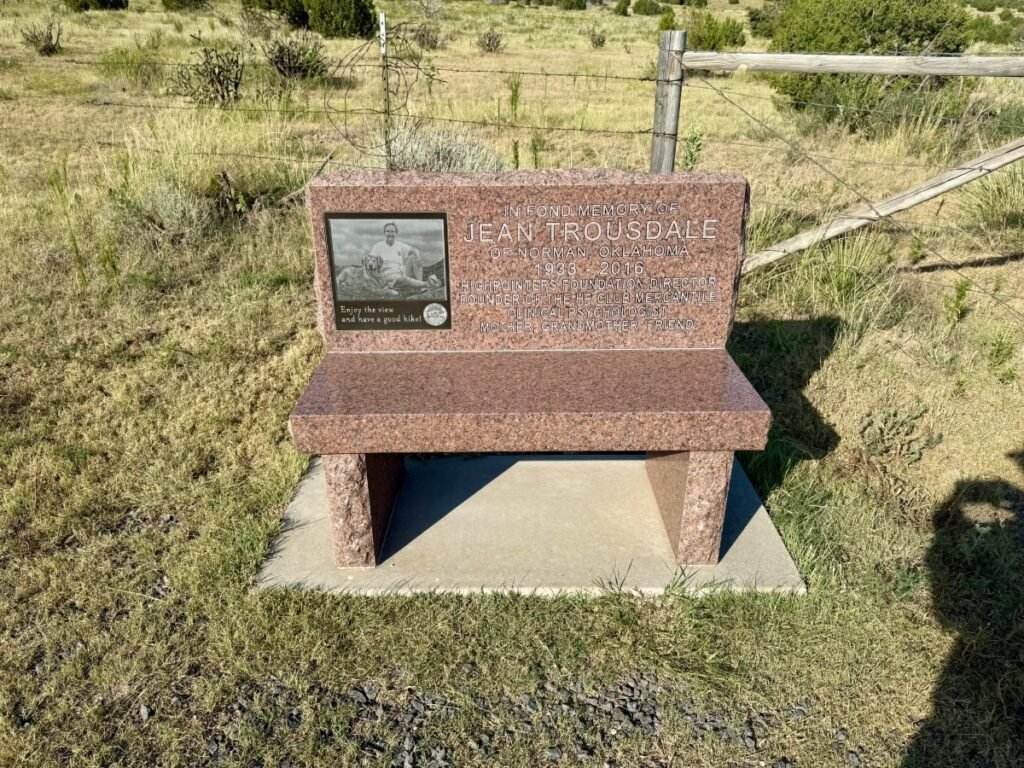 A stone memorial bench dedicated to Jean Trousdale at the Black Mesa trailhead, with an engraved photo and text, set against open grassland and scattered desert shrubs.