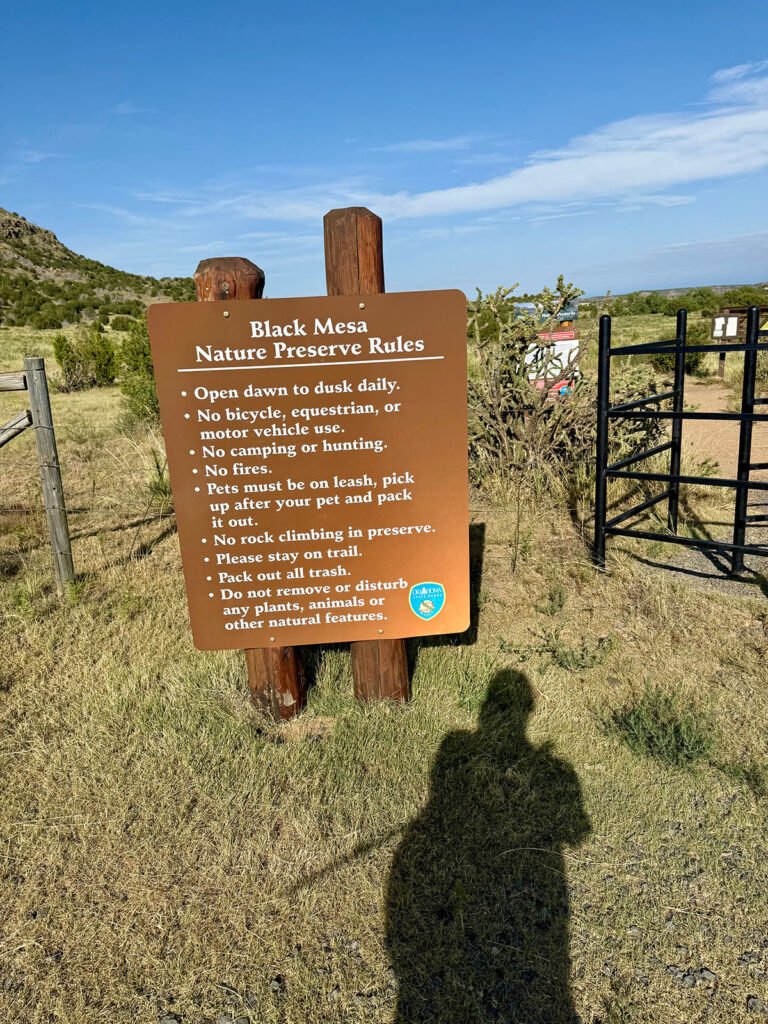 A brown wooden sign at the Black Mesa Nature Preserve trailhead listing preserve rules, with desert grass, shrubs, and rolling hills in the background under a clear blue sky.