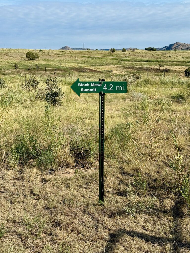 A green directional sign reading “Black Mesa Summit 4.2 mi.” stands in the middle of open grassland, with distant mesas and a partly cloudy sky in the background.