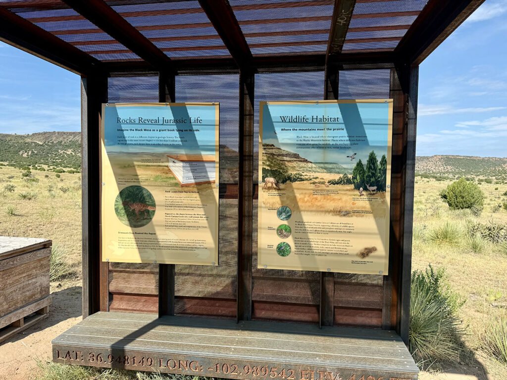 Interpretive panels at the Black Mesa hydration station describing Jurassic rock layers and local wildlife habitat, displayed in a shaded metal kiosk with desert grassland and mesas in the background.