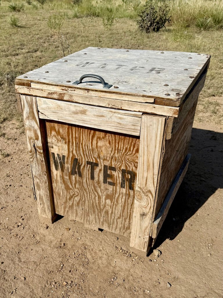 A weathered wooden box labeled “WATER” sitting along the Black Mesa trail, surrounded by dry dirt and desert grassland.