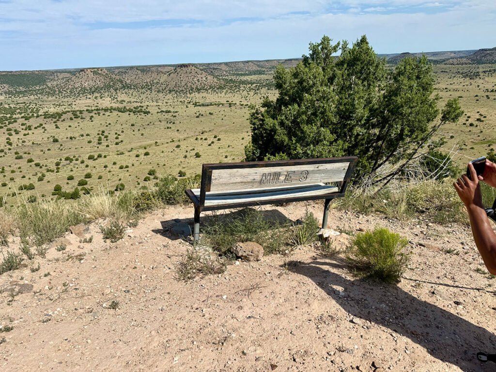 A wooden “Mile 3” bench perched along the Black Mesa trail overlooking a vast desert landscape, with a hiker’s arm visible holding a camera on the right side.