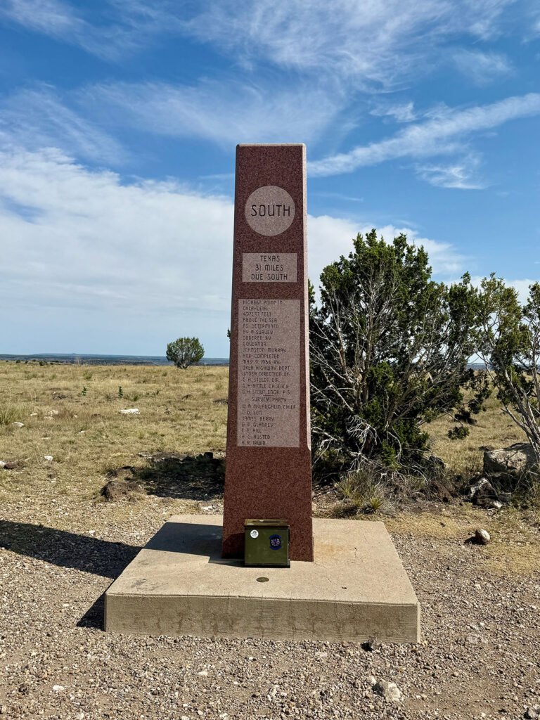 The “South” face of the Black Mesa summit obelisk, listing Texas at 3 miles due south, with low shrubs and wide-open plains behind it.