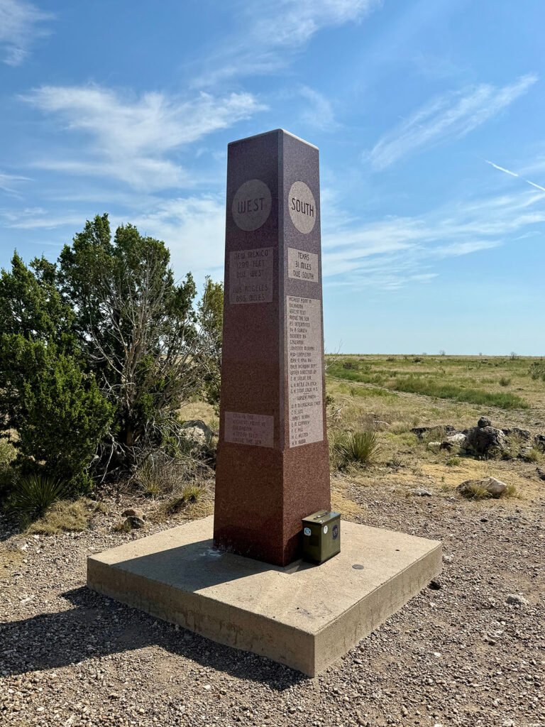 A tall red granite obelisk at the summit of Black Mesa showing the “West” and “South” directional markers, with distances to New Mexico and Texas, standing on a concrete base in open grassland.