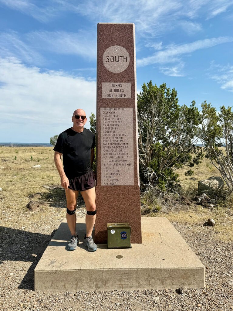 A hiker standing next to the Black Mesa summit obelisk, with the green metal register box and the round USGS benchmark visible on the concrete base beneath the monument.