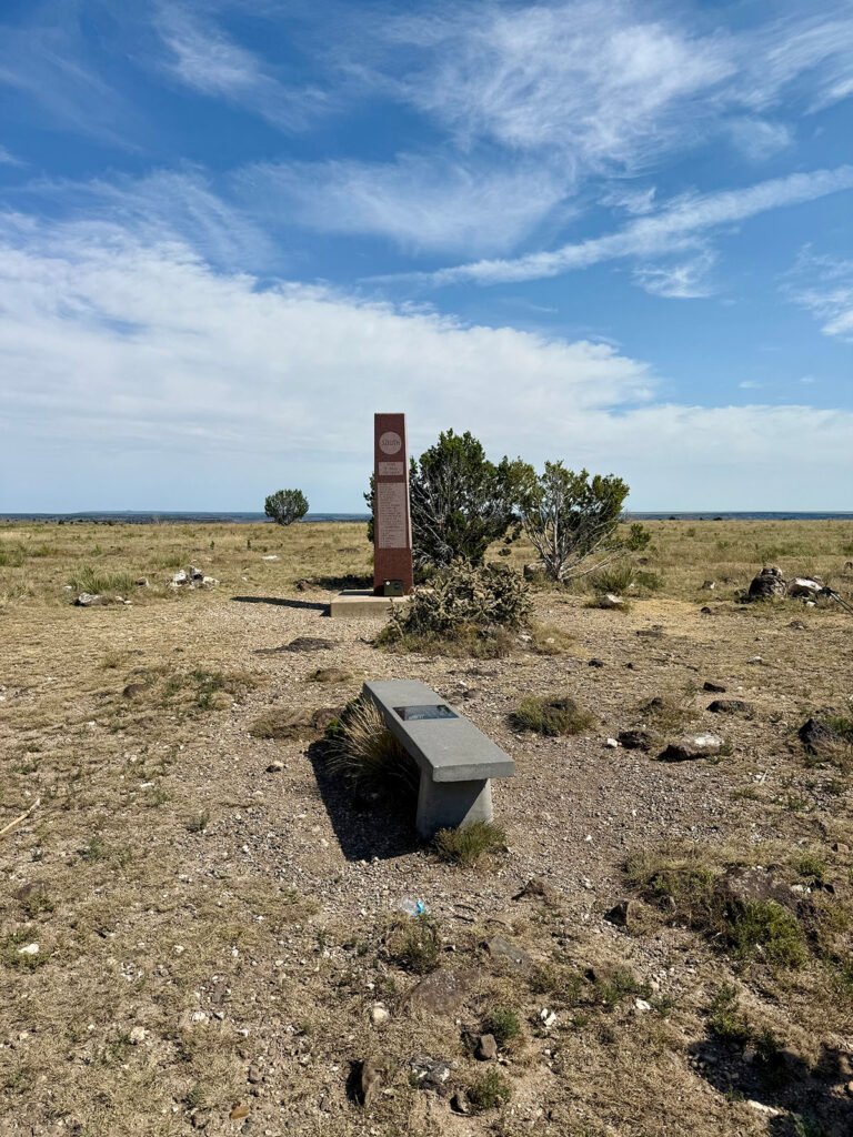 A wide view of the summit area on Black Mesa with a stone bench in the foreground and the red granite summit obelisk in the distance under a blue sky.