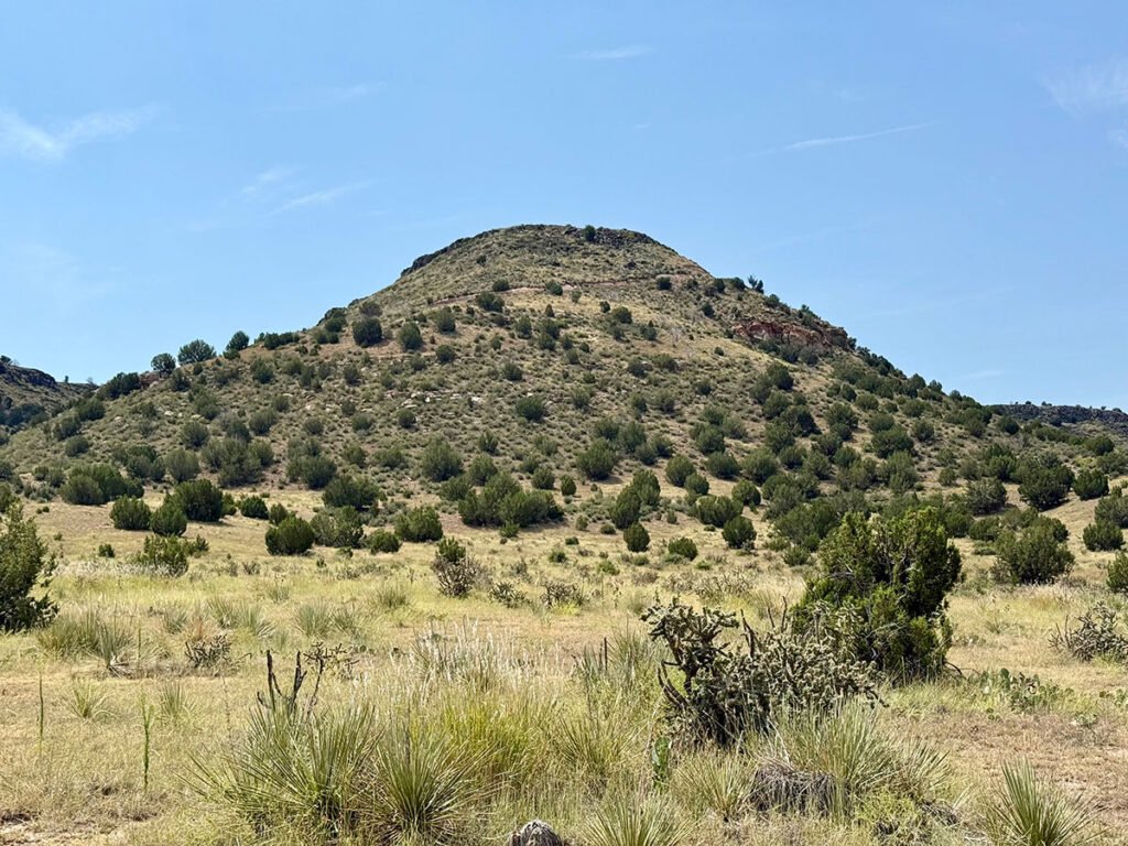 A view from the base of Black Mesa looking upward, with a faint hiking trail visible etched across the slope among scattered shrubs and desert vegetation.