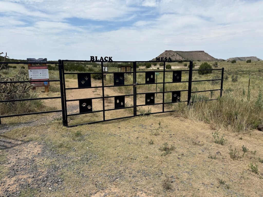 A black metal gate marking the entrance to the Black Mesa trailhead, decorated with cut-out animal tracks, with mesas and wide desert landscape stretching out behind it.