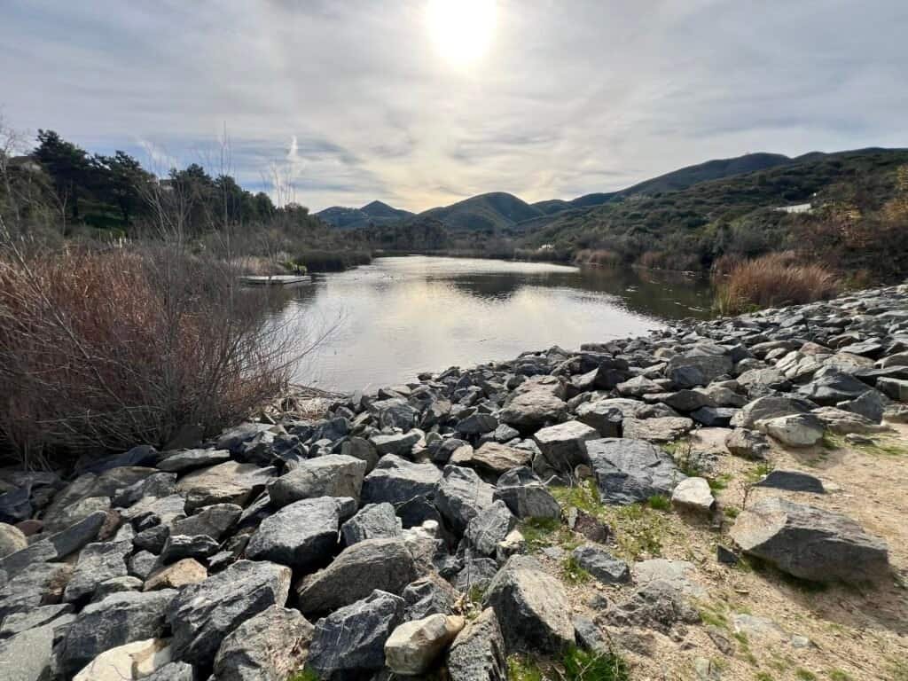 Rock-lined shoreline along Discovery Lake with calm water reflecting surrounding hills in San Marcos, California.