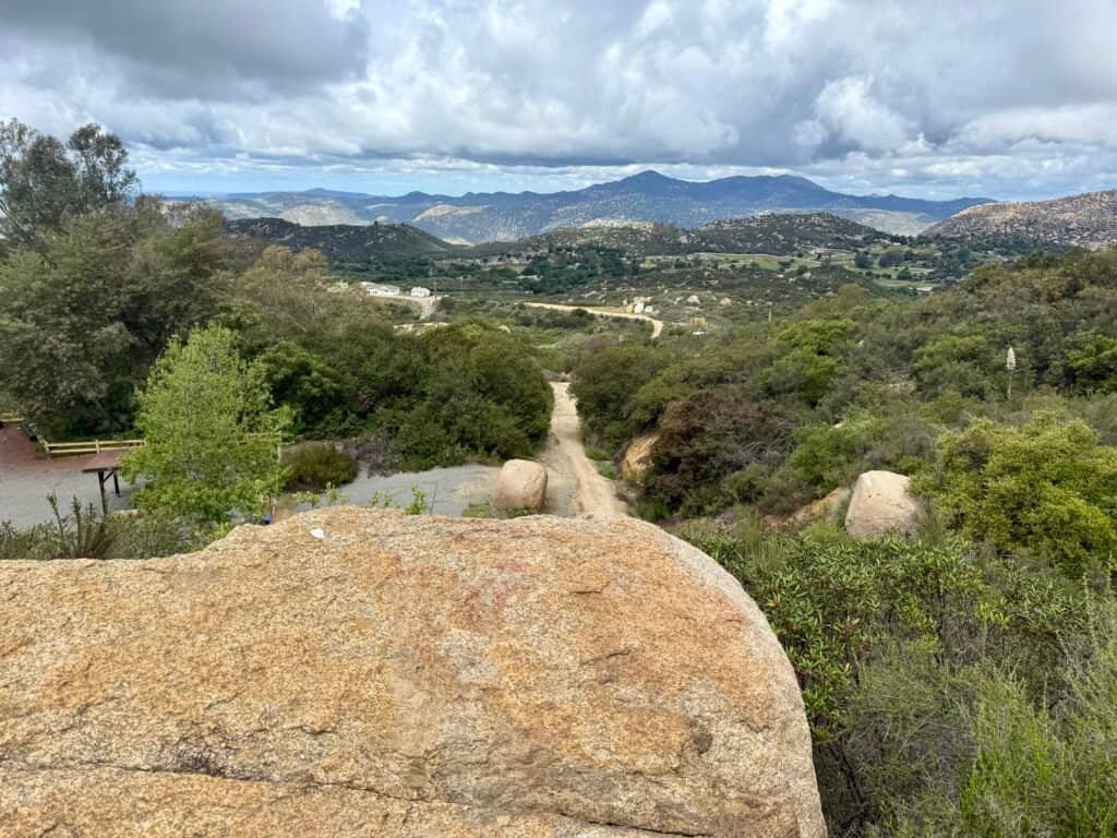 Looking back down the El Cajon Mountain trail toward the trailhead, with rolling hills, trees, and storm clouds overhead.
