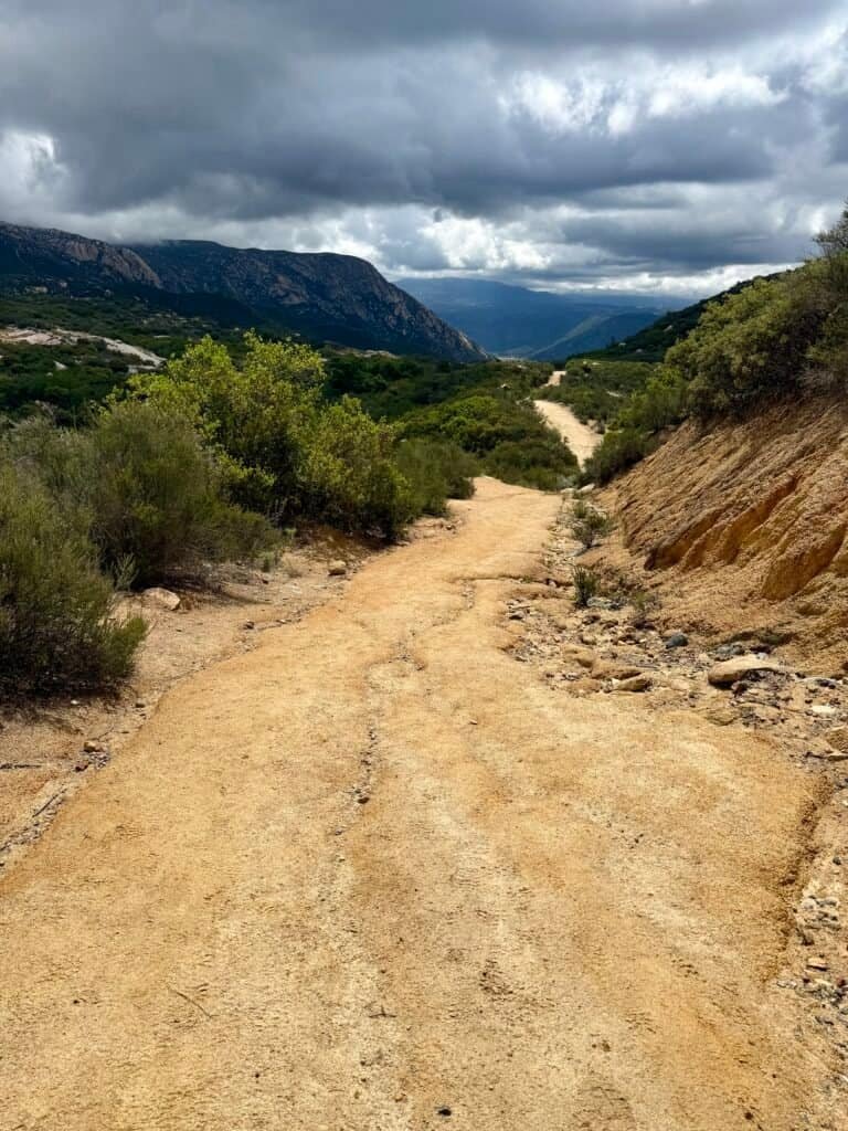Looking back down the winding trail on El Cajon Mountain under heavy clouds, with rolling hills and distant ridgelines.