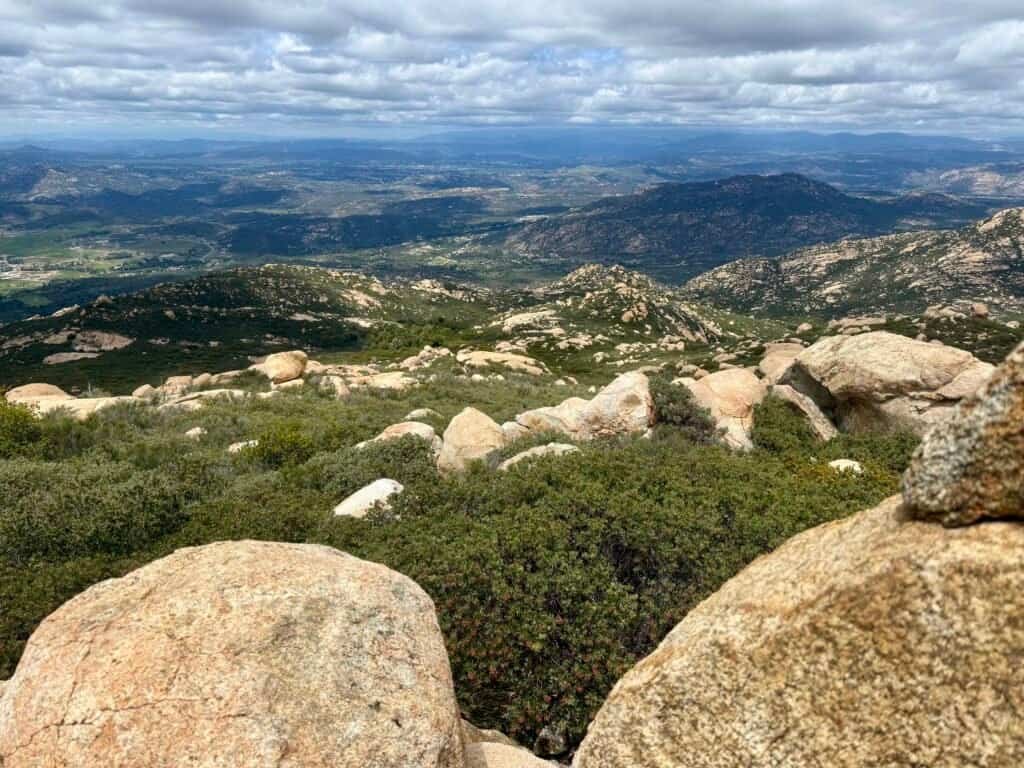 Wide view from El Cajon Mountain summit showing rocky terrain, rolling hills, and distant valleys under layered clouds.