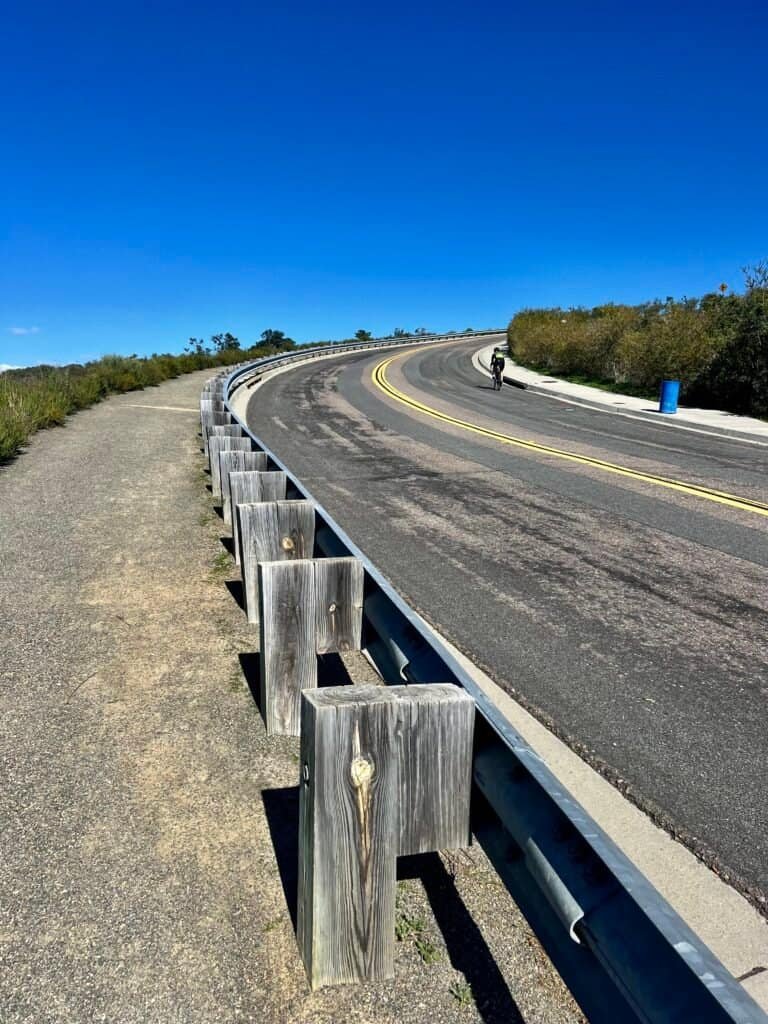Hiker walking along the paved shoulder of Double Peak Drive in San Marcos, California, with a curving road, guardrail, and clear blue sky leading toward the summit.