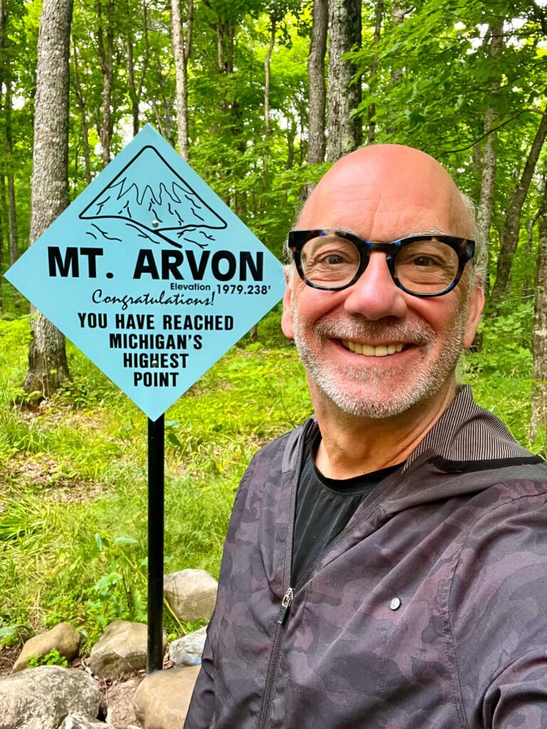 Smiling hiker standing beside the Mt. Arvon summit sign marking Michigan’s highest point at 1,979 feet in the Upper Peninsula.