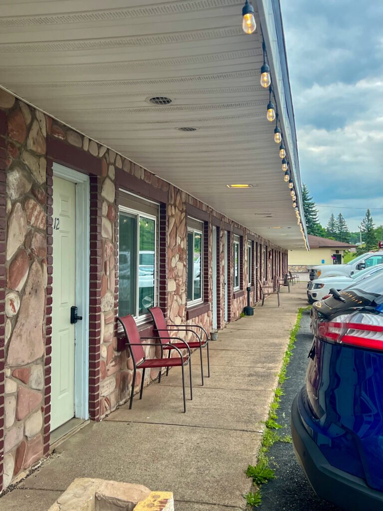 Exterior corridor of Motel 41 in L’Anse, Michigan with stone façade rooms, red chairs, and parked cars under string lights.