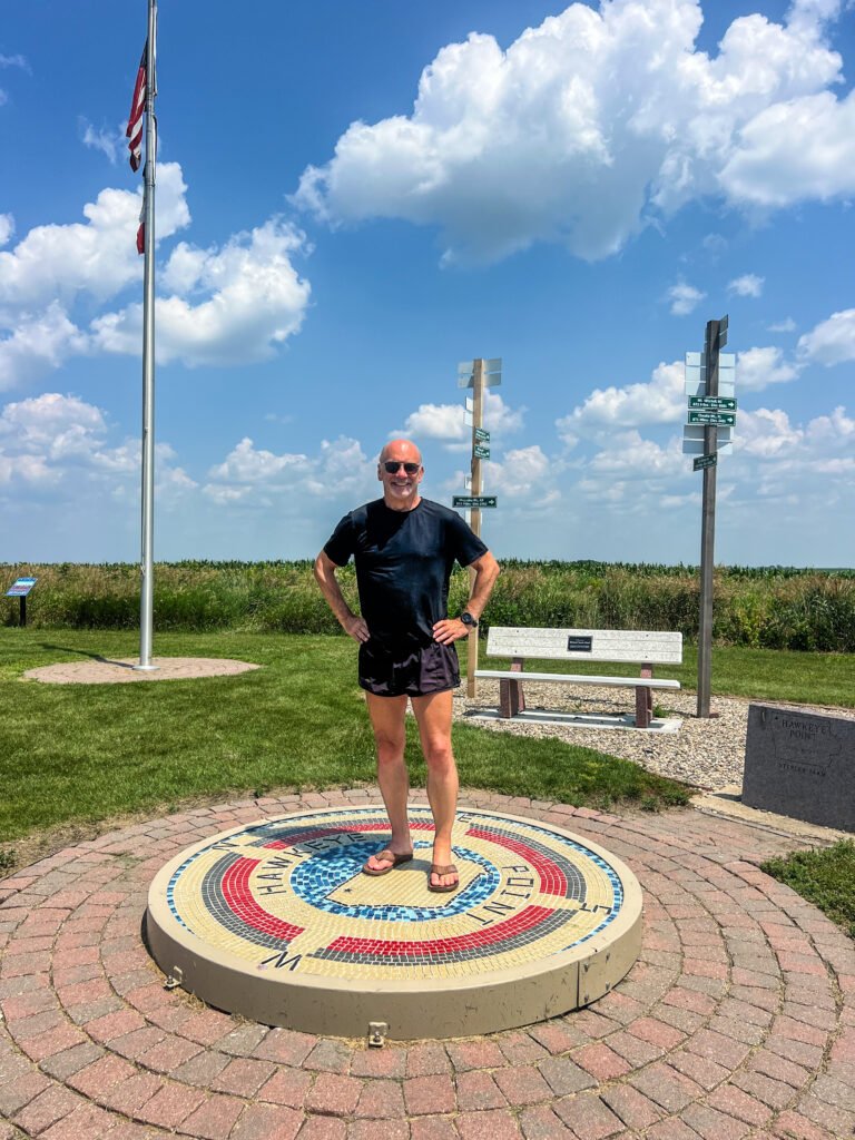 Stephen standing on the Hawkeye Point summit mosaic in Iowa with directional signs, flagpole, and cornfields behind him.