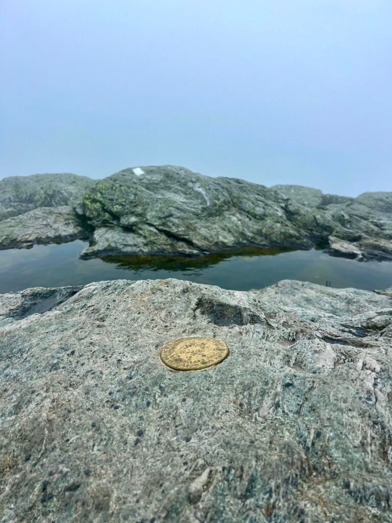 USGS summit marker surrounded by fog and puddled rock at the top of Mount Mansfield.
