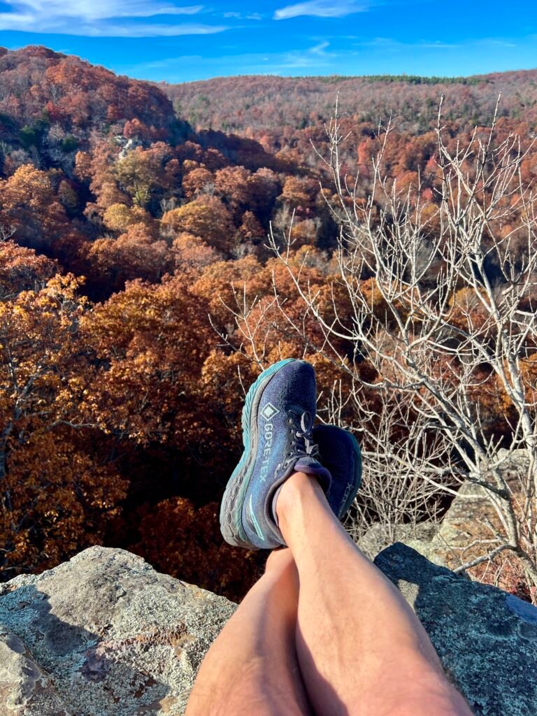 Blue hiking shoes resting on a rocky cliff at Inspiration Point, overlooking colorful fall foliage in Mount Magazine State Park, Arkansas.