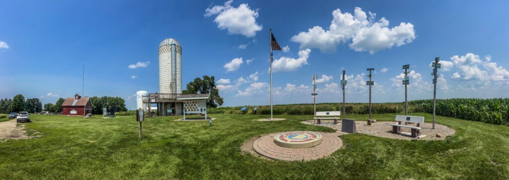 Wide panoramic view of Hawkeye Point summit area with silo, red barn museum, summit mosaic, flagpole, benches, and cornfields in northwest Iowa.