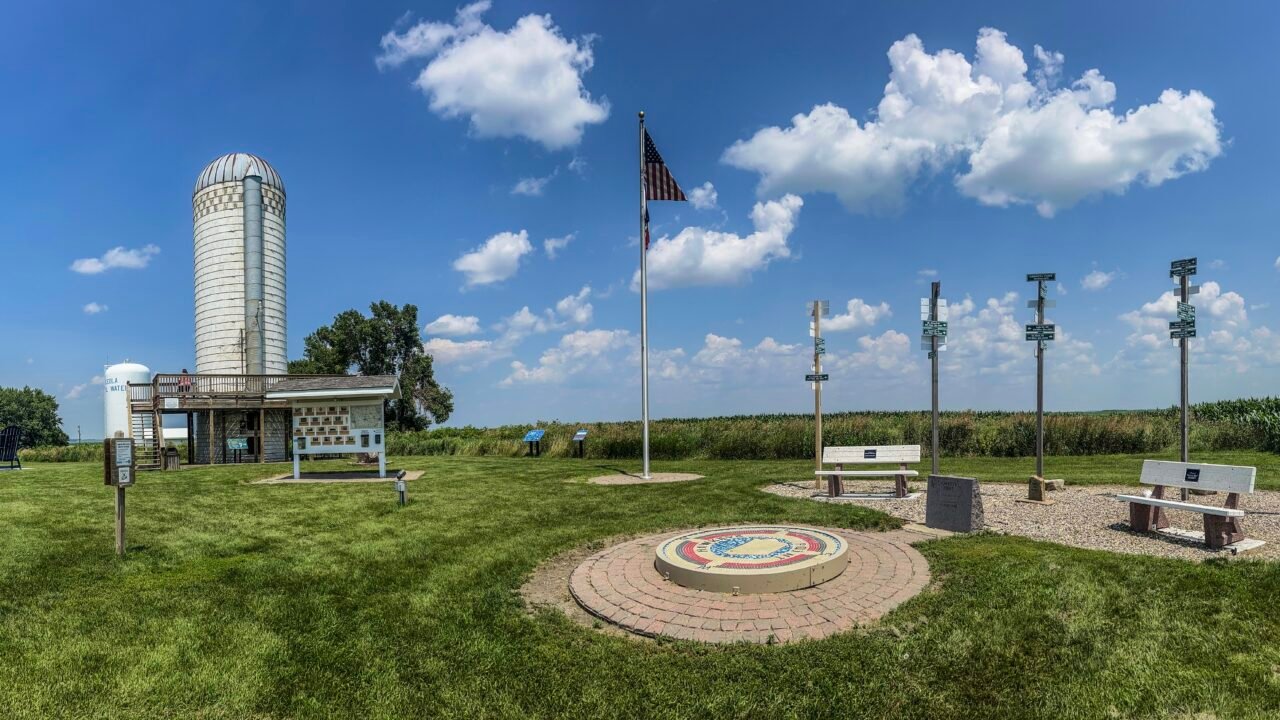 Panoramic view of Hawkeye Point summit in Iowa featuring the high point mosaic, American flag, silo observation deck, benches, and surrounding cornfields under a blue sky.