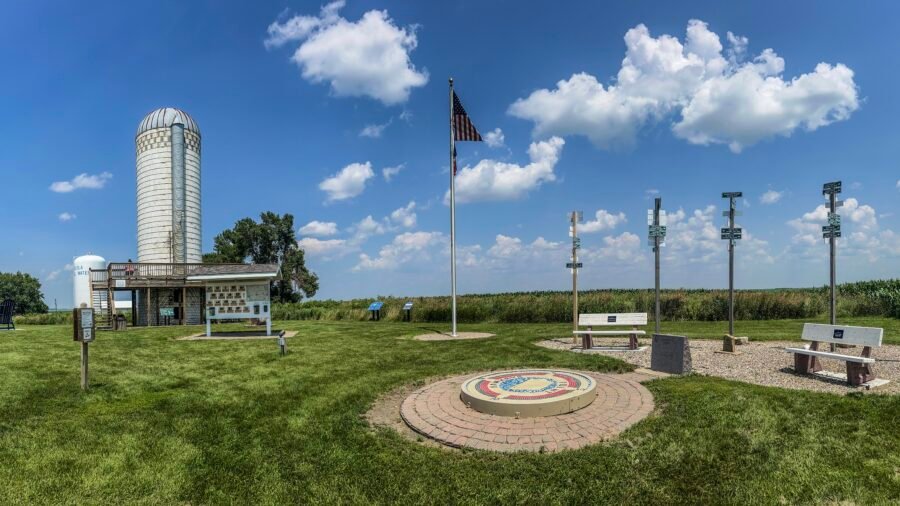 Panoramic view of Hawkeye Point summit in Iowa featuring the high point mosaic, American flag, silo observation deck, benches, and surrounding cornfields under a blue sky.