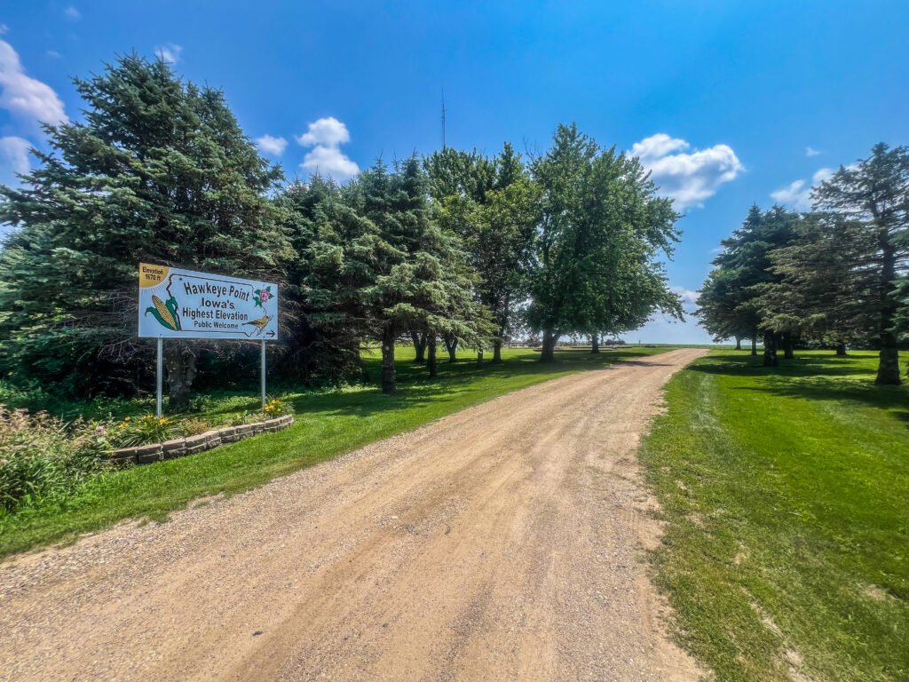Gravel road lined with trees leading to Hawkeye Point summit area in rural northwest Iowa under a blue sky.